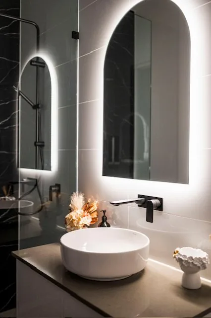 Walk in shower beside a floating vanity with backlit arch mirror