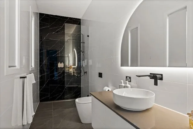 Black marble shower recess beside a backlit arch mirror and stone top